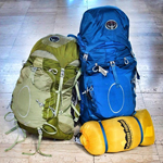 Large blue Osprey backpack supporting a smaller green Osprey backpack with a small yellow draw string pack laying on tile in front of both. 