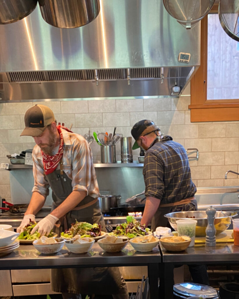 One chef with their back turned toward us is sautéing on the range while another chef, facing us is busy plating dishes for guests while working a destination job. 