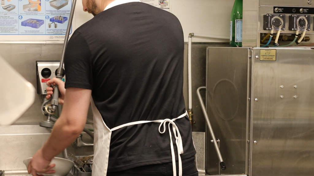 A dishwasher wearing a black t-shirt and a white apron in the dish pit spraying off dirty plates to put in the dishwasher. 